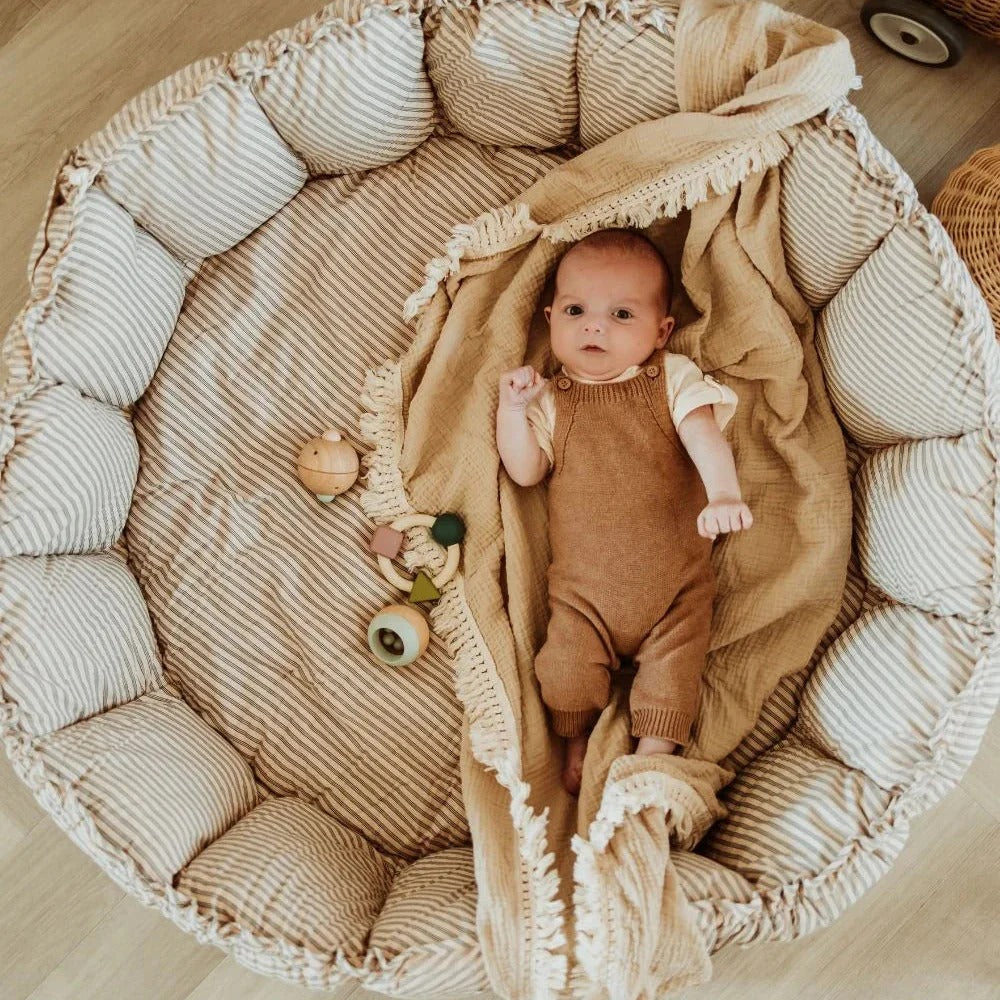Baby in a textured basket with a blanket, surrounded by toys on a wooden floor.