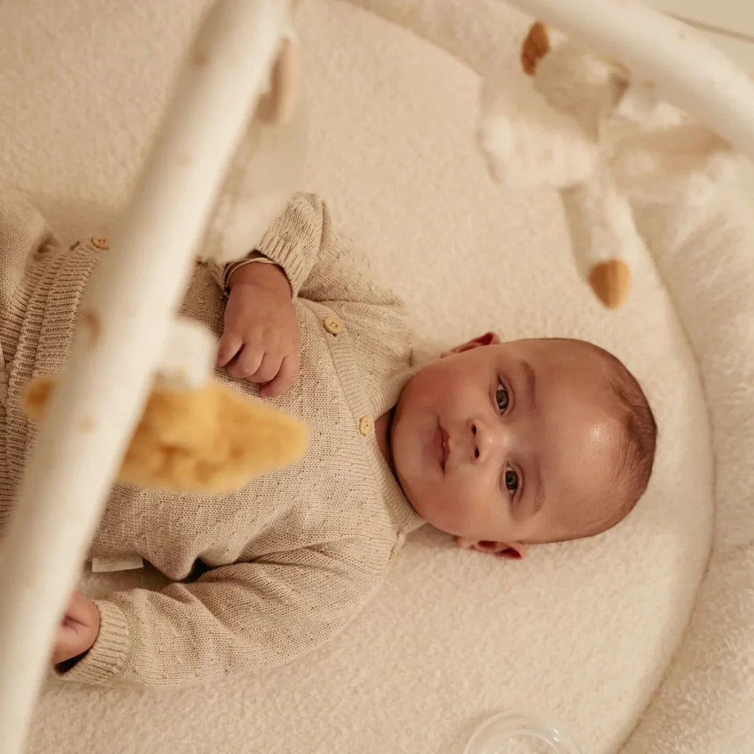 Baby lying in a crib wearing a beige outfit with teddy bear toys.