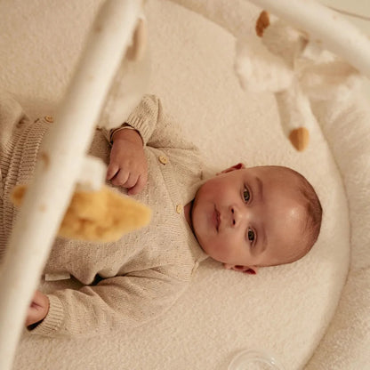 Baby lying in a crib wearing a beige outfit with teddy bear toys.