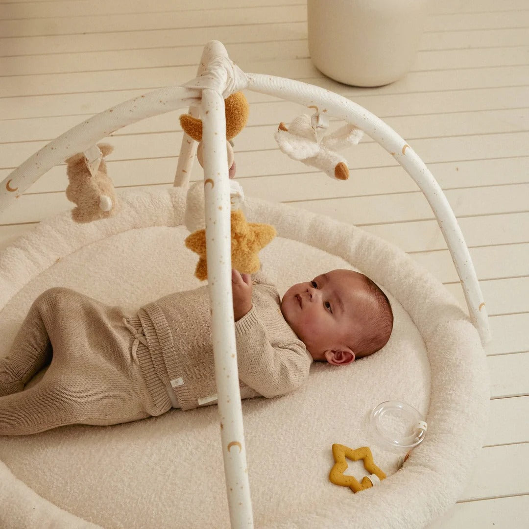 Baby lying in a white crib with toys, wearing a beige outfit.