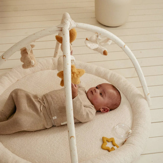 Baby lying in a white crib with toys, wearing a beige outfit.