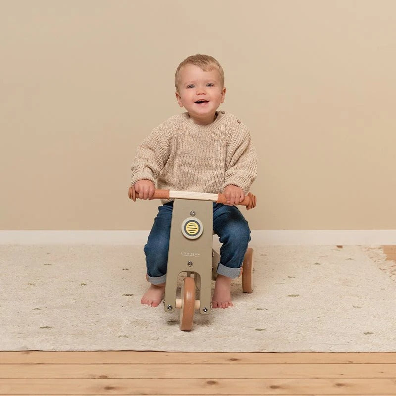 Child riding a wooden balance bike on a wooden floor with a beige wall background