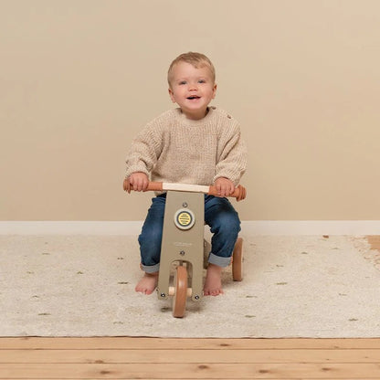 Child riding a wooden balance bike on a wooden floor with a beige wall background