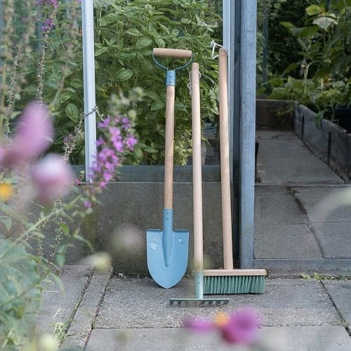 Gardening tools including a spade, rake, and broom leaning against a wall with flowers and plants in the foreground.