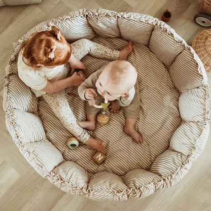 Two babies playing in a round, cushioned playpen on a wooden floor.