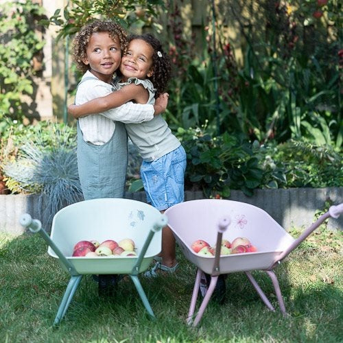 Two children hugging in a garden with two wheelbarrows filled with apples.
